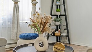 Un salon avec une table ronde en bois, un vase blanc contenant des plantes séchées, un coussin bleu et une étagère noire en forme d’échelle, ornée de plantes.