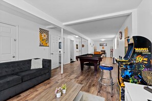 A modern living room with a pool table, a black sofa, and a vibrant Marvel-themed arcade machine.
