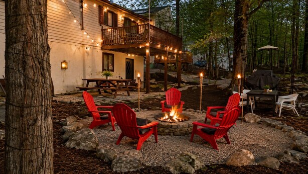 Un espace extérieur rustique aménagé avec un foyer, des fauteuils Adirondack rouges et une terrasse en bois illuminée de guirlandes lumineuses.