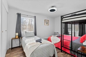 Une chambre avec un lit superposé, une table de chevet avec une lampe, une fenêtre avec des rideaux et un ventilateur de plafond.
