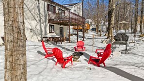 Un espace extérieur aménagé avec des sièges, recouvert de neige, comprenant des chaises rouges et un foyer, le tout entouré d’arbres et d’une maison dotée d’un balcon.
