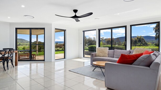 A spacious living room with a grey sofa, a wooden coffee table, and a dining area with a table and chairs. Large windows offer a view of a green landscape and hills. - Snowy Mountains Retreat - 10 Mowamba Way (Moonbah)