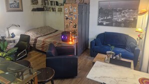 En kompakt opholdsstue med seng, blå sofa, lille bord, skrivebord med lampe og bogreol.