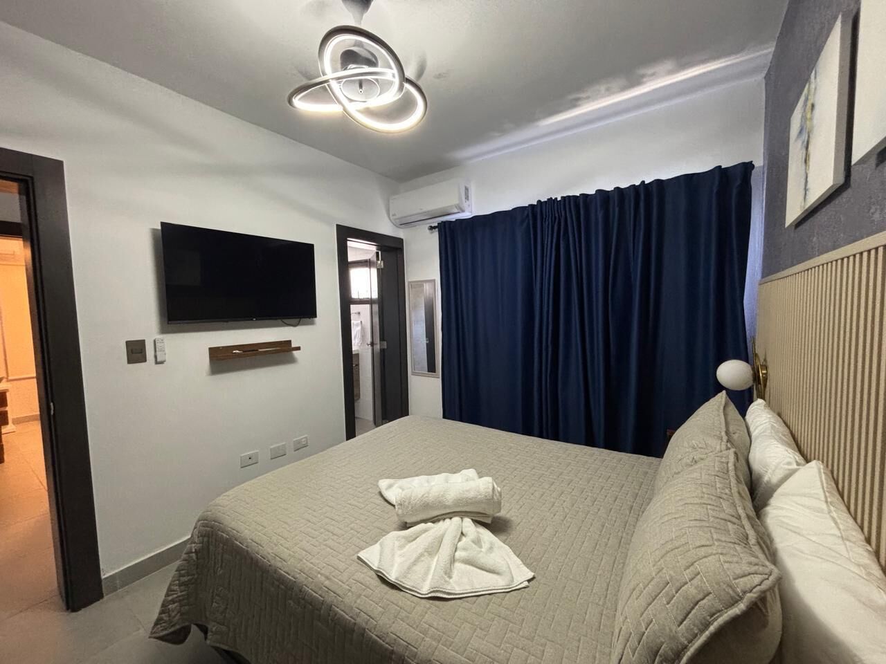 A hotel room with a bed, a flat-screen TV mounted on the wall, a ceiling light fixture, and a window with curtains.
