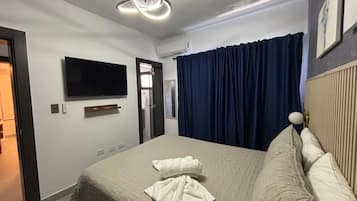 A hotel room with a bed, a flat-screen TV mounted on the wall, a ceiling light fixture, and a window with curtains.