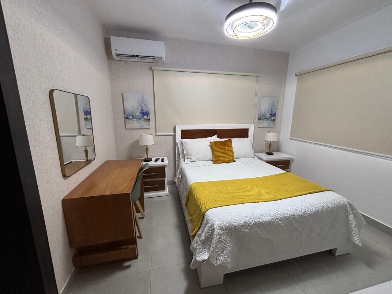 Una habitación de hotel con cama, escritorio, silla, mesita de noche, lámpara y aire acondicionado instalado en la pared.