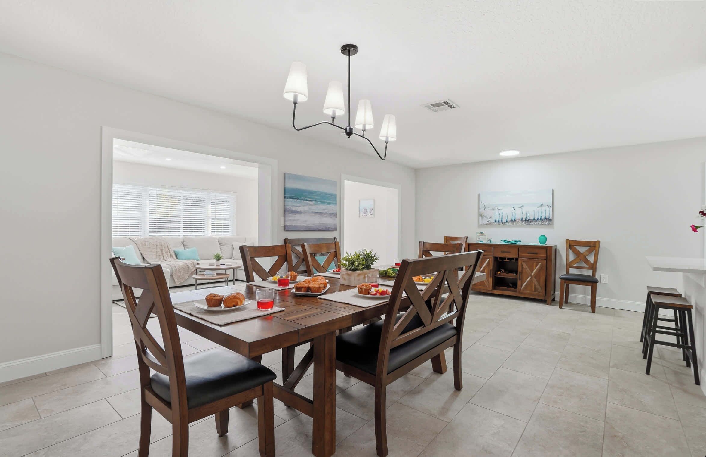 A dining area with a wooden table, chairs, and a chandelier. There is a sofa and a cabinet in the background.