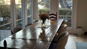 Un'area pranzo con un tavolo e sedie in legno, un vaso con fiori e ampie finestre vista alberi e un edificio.
