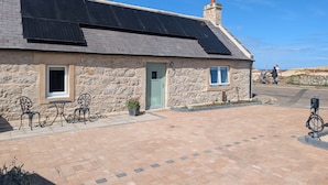 A stone building with a green door, solar panels on the roof, and a paved area with outdoor furniture. - Warm and accessible coastal cottage with sustainable design — coming soon (Lossiemouth)