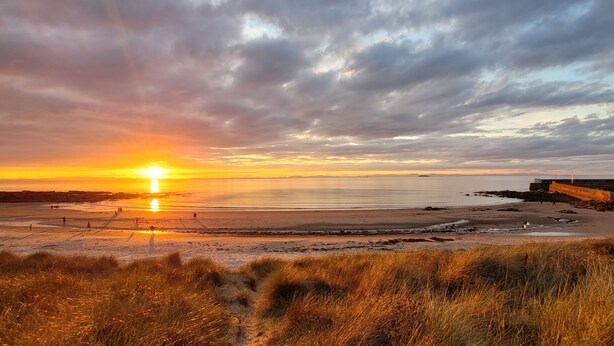 A beach with a sunset, grassy dunes, and a distant shoreline. - Warm and accessible coastal cottage with sustainable design — coming soon (Lossiemouth)