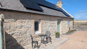 A stone building with a tiled patio, a chimney, and solar panels on the roof. - Warm and accessible coastal cottage with sustainable design — coming soon (Lossiemouth)