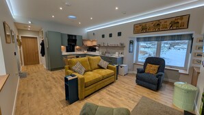 A modern living room with a sofa, armchair, and a dining area with a kitchen in the background. - Warm and accessible coastal cottage with sustainable design — coming soon (Lossiemouth)