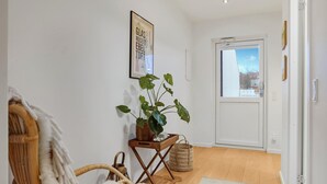 Un couloir doté d’un plancher en bois, d’une chaise en osier, d’un tapis blanc et moelleux, d’une plante en pot et d’une porte donnant sur l’extérieur.