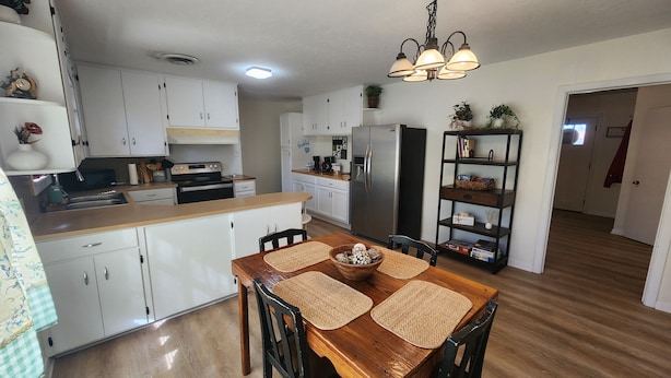 A kitchen with white cabinets, a wooden dining table with woven placemats, a stainless steel refrigerator, and a hanging light fixture. - The Brenham Abode is pet friendly and conveniently located close to downtown. (Brenham)