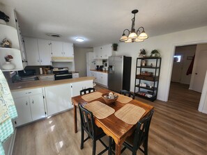 Una cocina con armarios blancos, una mesa de comedor de madera con individuales tejidos, un refrigerador de acero inoxidable y una lámpara colgante.