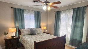 A bedroom with a wooden bed, two bedside tables with lamps, a ceiling fan, and large windows with curtains. - The Brenham Abode is pet friendly and conveniently located close to downtown. (Brenham)