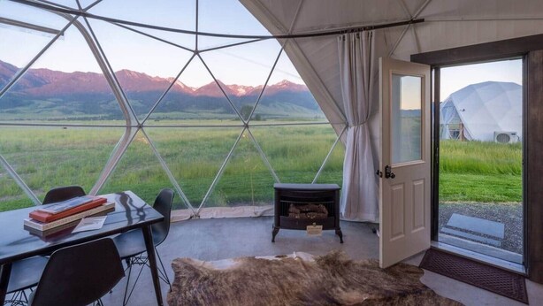 Uma tenda com teto de vidro, uma mesa com livros, uma cadeira, uma pequena mesa com lareira e vista para montanhas.