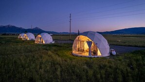 A row of dome-shaped tents with interior lighting, set in a grassy field with mountains in the background. - Yellowstone Luxury Dome w/ Views & Stargazing (Park County)