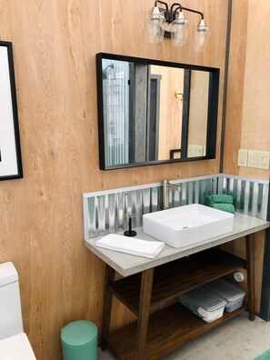 A bathroom with a wooden wall, a white sink with a mirror above it, and a green trash bin.