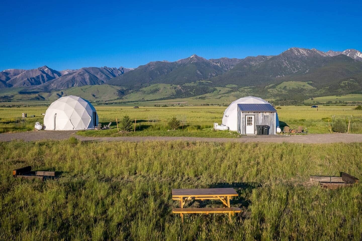 Dos edificios con forma de cúpula en un campo cubierto de hierba, con montañas al fondo.