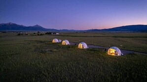 Quatre tentes en forme de dôme, illuminées au crépuscule, sont installées dans un vaste champ ouvert, avec des montagnes en toile de fond.