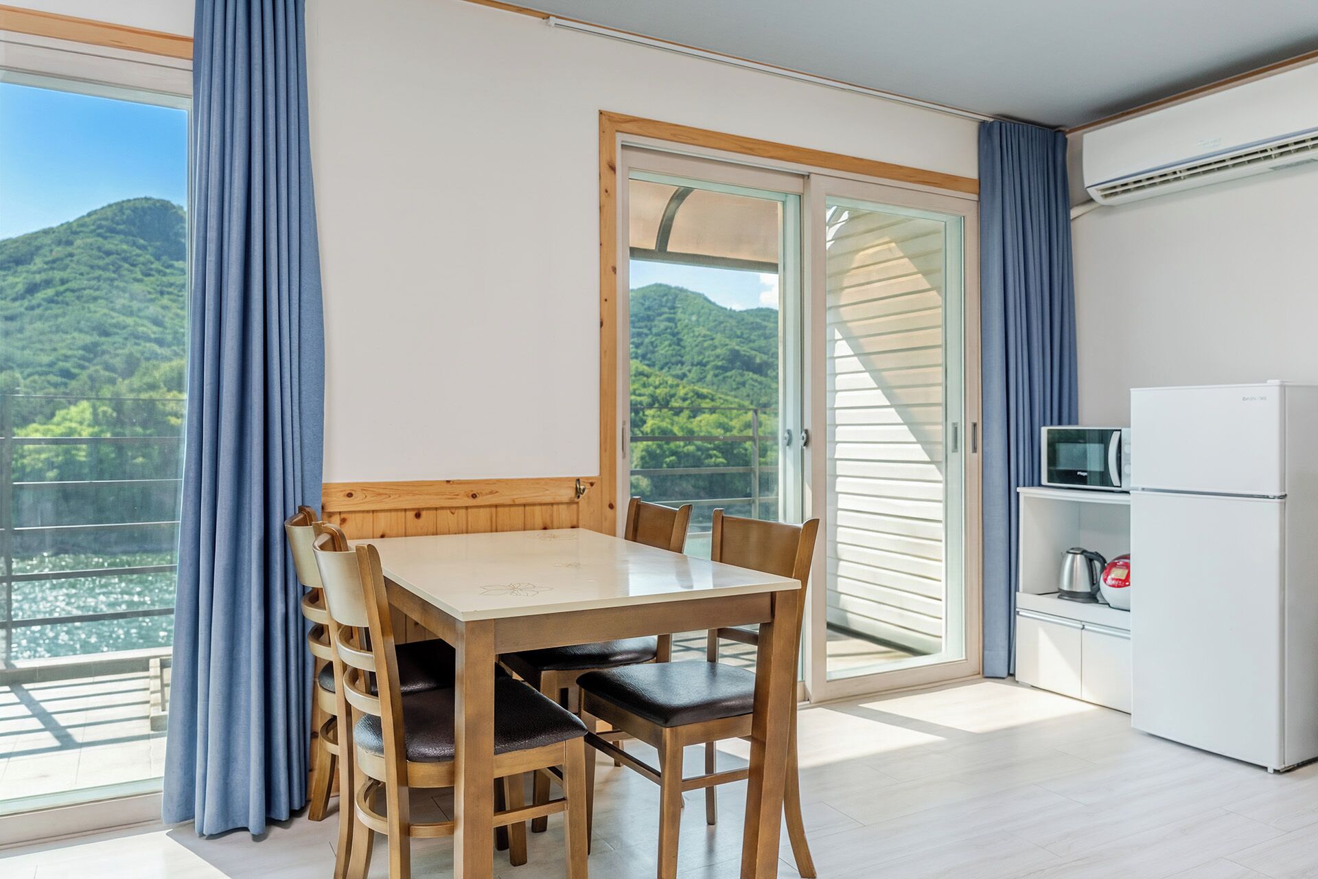 Una habitación con una mesa de comedor, sillas, una zona de cocina y un ventanal corredero que da a un balcón con vistas a una zona boscosa.
