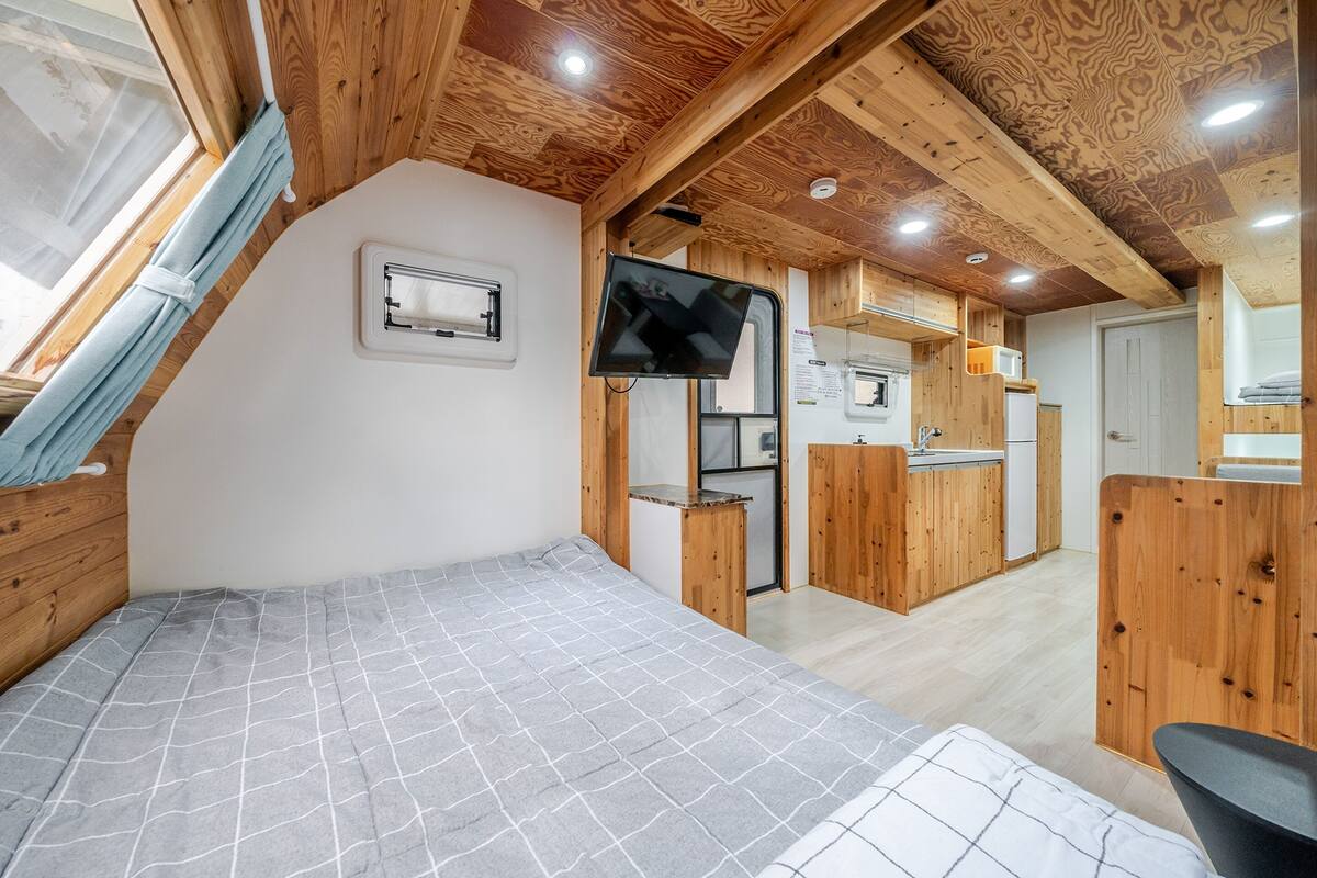 A compact room with a bed, a TV mounted on the wall, a small kitchenette, and a window with a curtain.