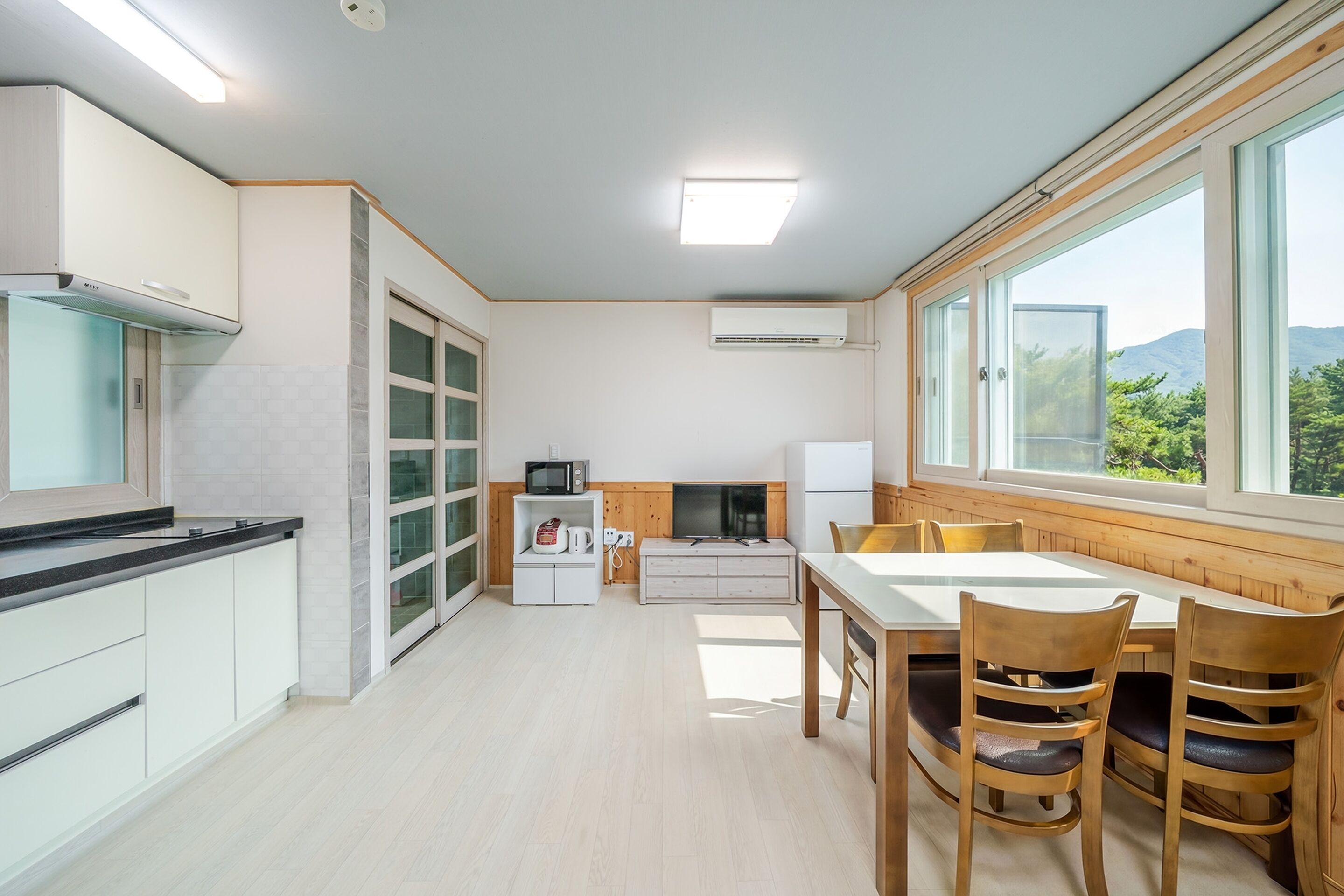 A modern kitchen with a dining area, large windows, and a view of greenery.