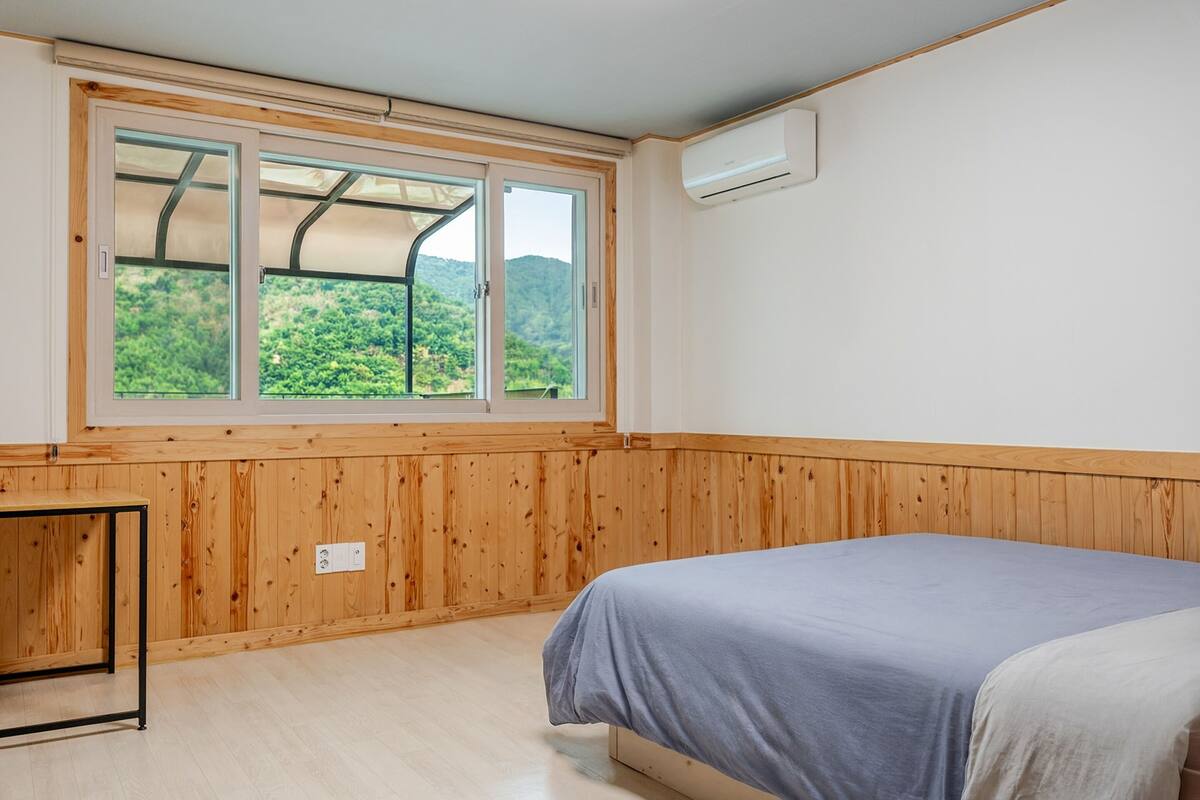 A room with a bed, wooden paneling, a window with a view of greenery, and an air conditioning unit.