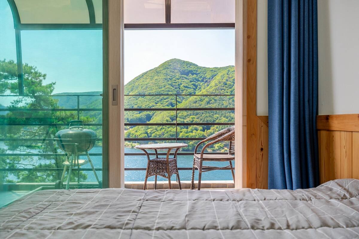 A bedroom with a large window, a bed, and a view of a mountain and lake.