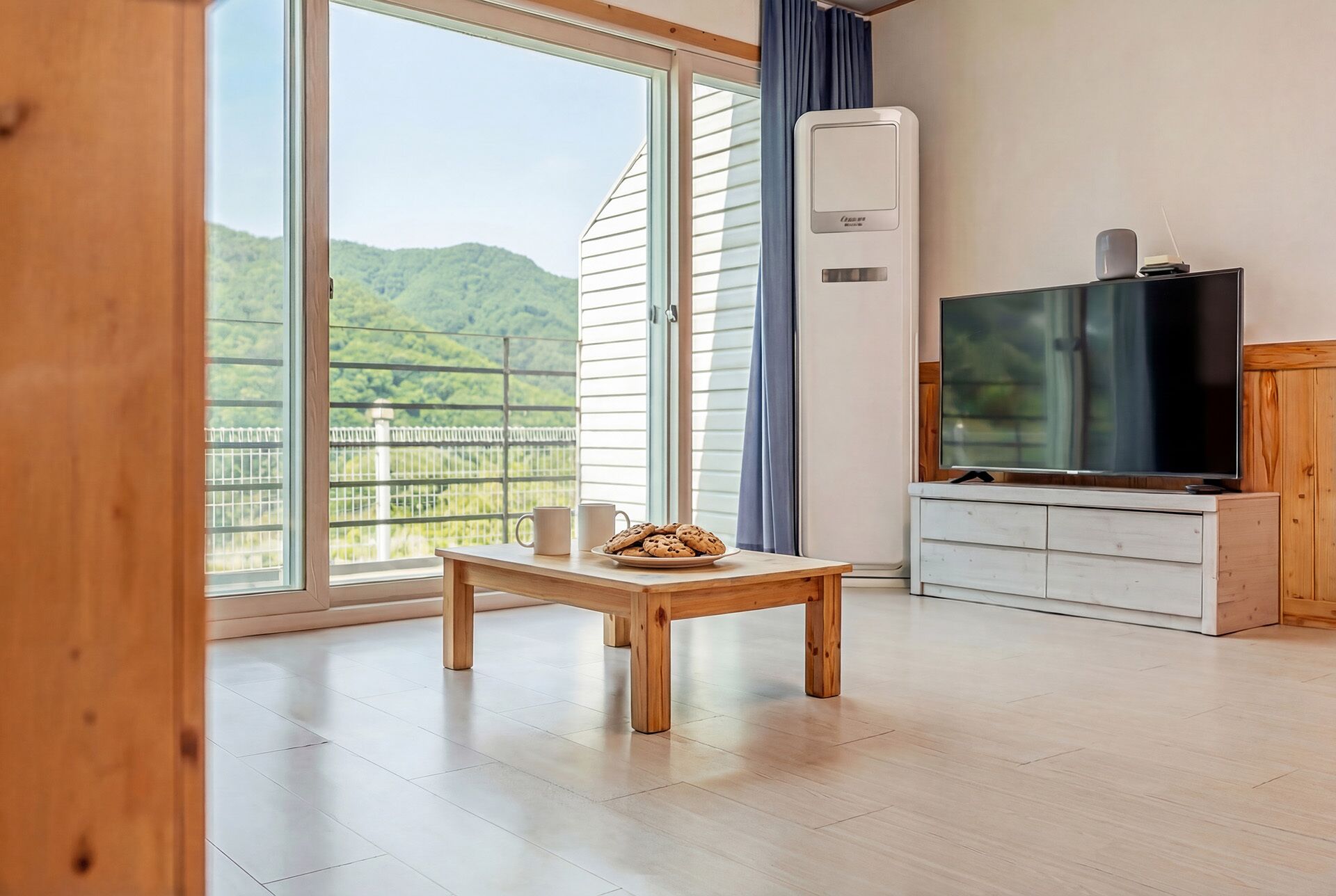 A room with a wooden table, a television, and a large window with a view of greenery.