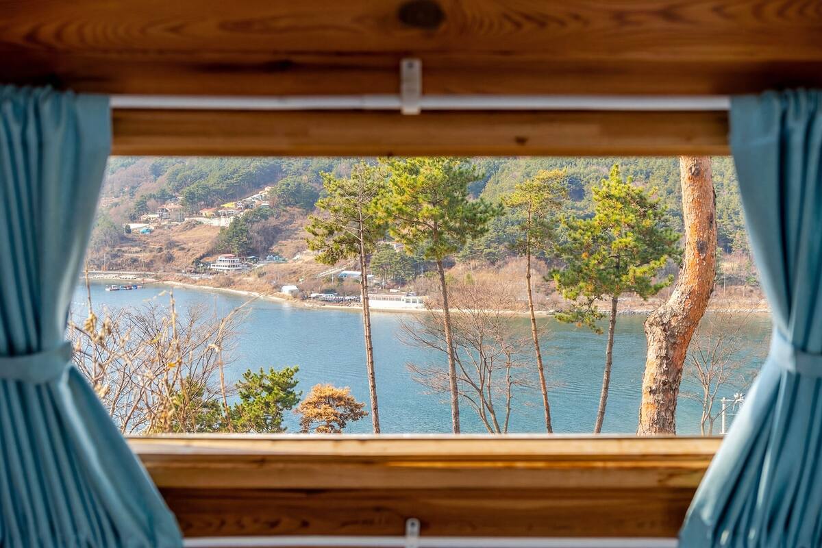 A cabin with a large window overlooking a lake and forested hills.