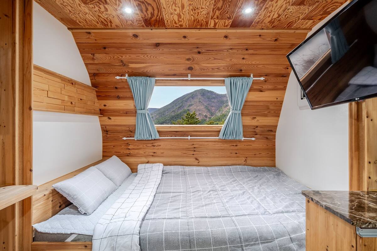 A bedroom with a wooden ceiling, a bed with a grey checkered duvet, and a window with curtains.
