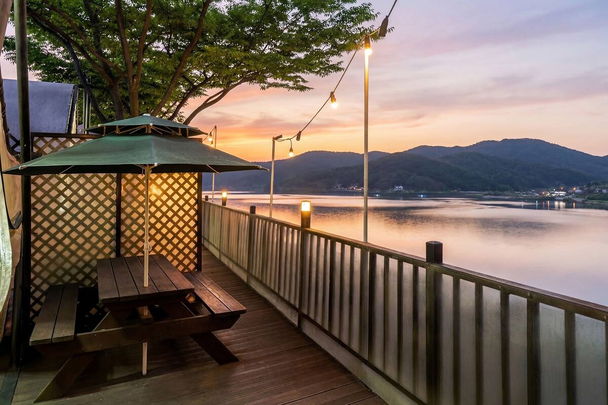 A wooden deck with a picnic table and a covered seating area, overlooking a calm body of water and distant mountains.
