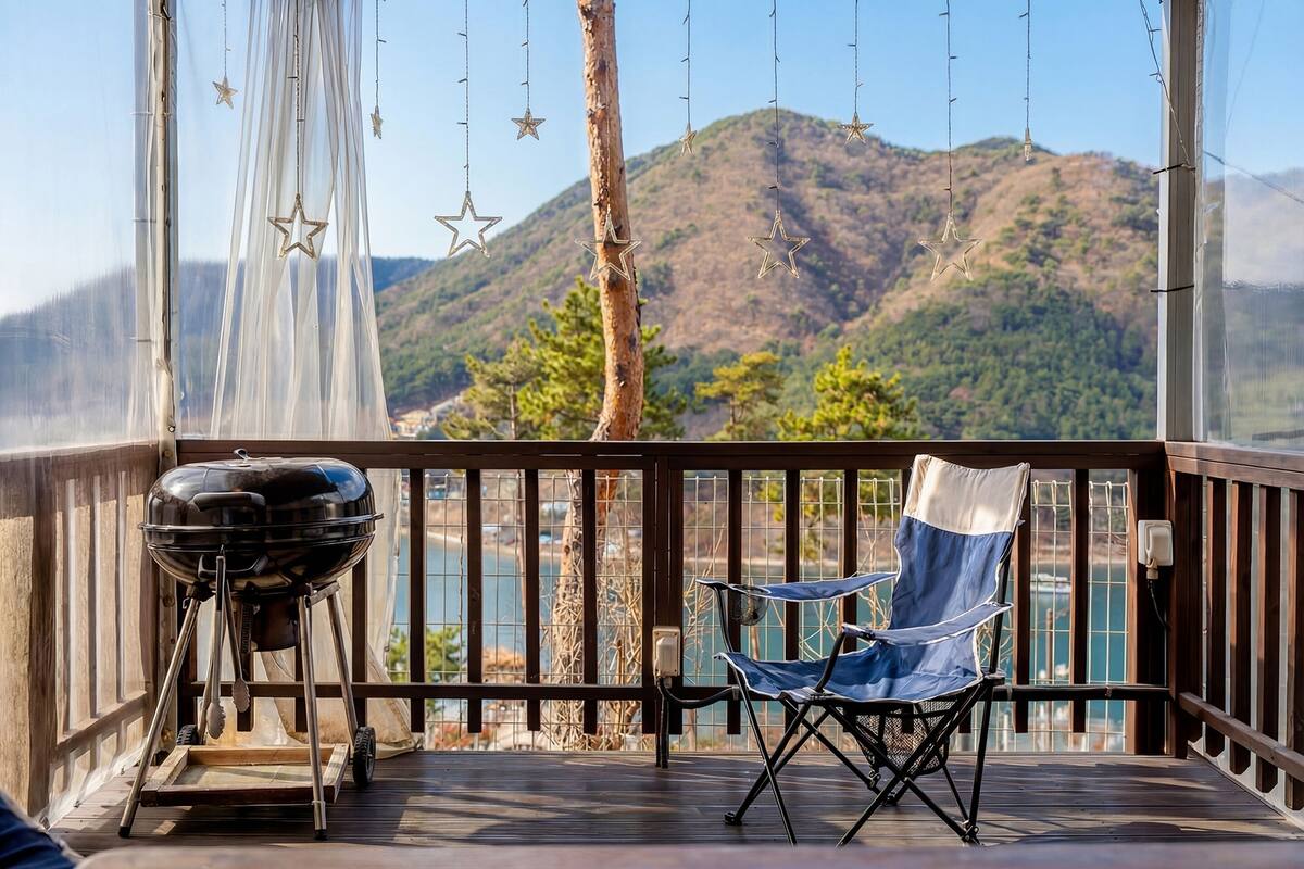 A balcony with a grill, a folding chair, and a view of a mountain and lake.