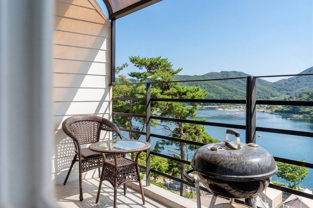 A balcony with wicker chairs, a glass table, and a barbecue grill overlooking a lake and mountains.