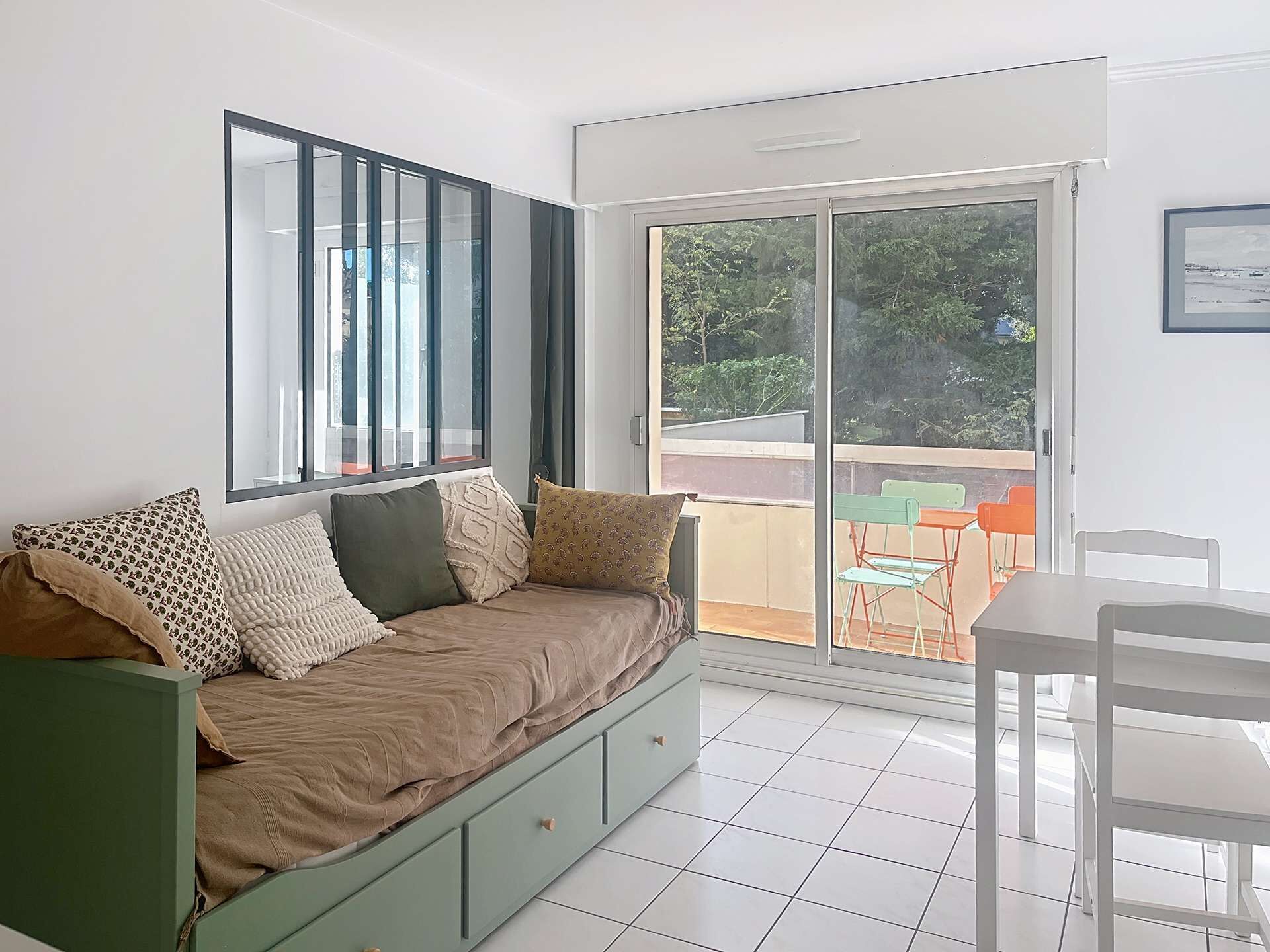 Een kamer met een groene bank, een witte tafel en een glazen schuifdeur die leidt naar een buitenruimte met kleurrijke stoelen.