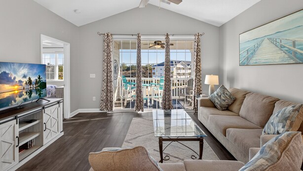 A spacious living room with a large window, a ceiling fan, a TV, and a sofa. - Ironwood | Pool & Tennis Near the Beach (North Myrtle Beach)