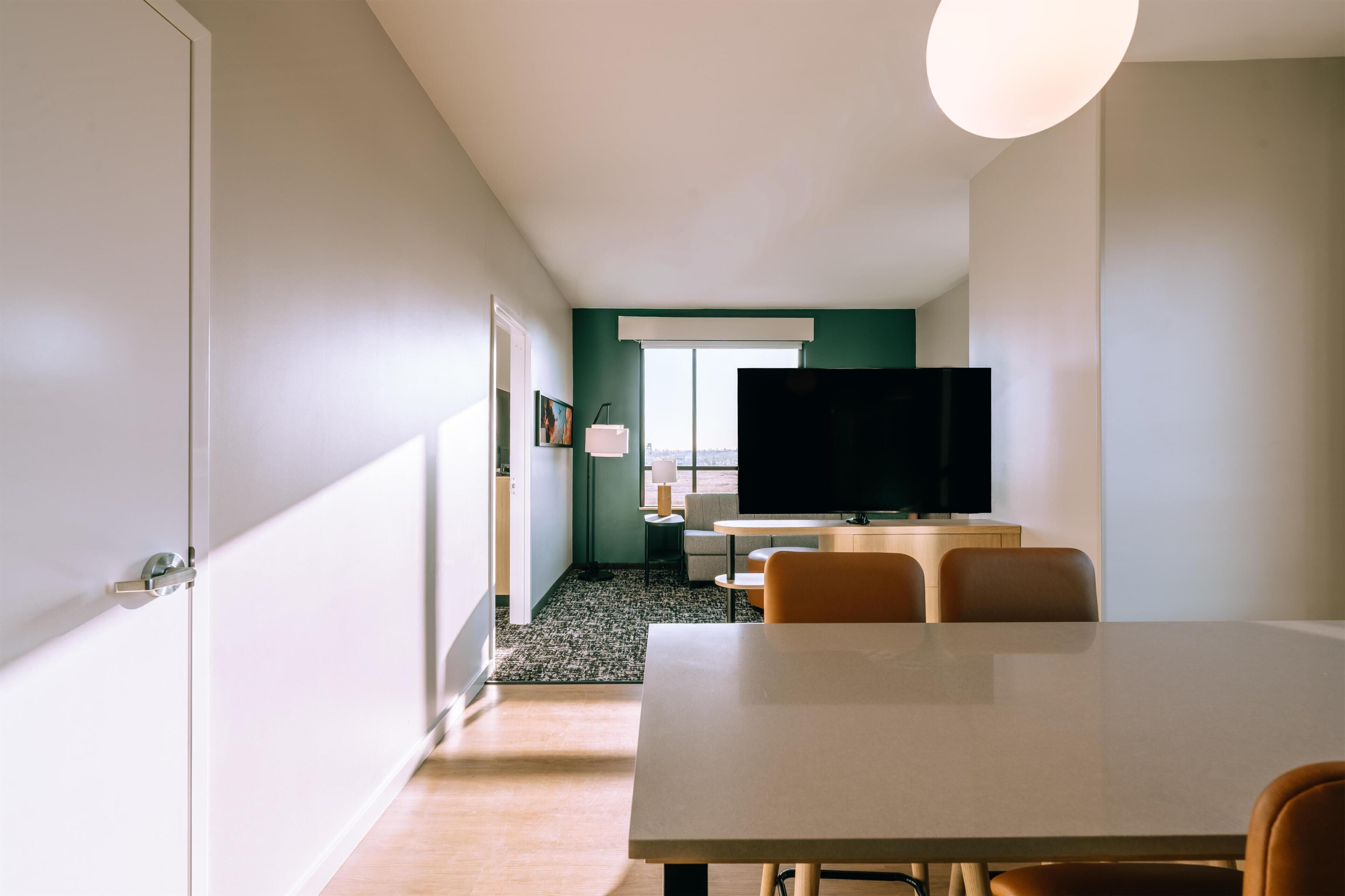A modern hotel room with a large bed, a flat-screen TV, a dining table, and a view of the outdoors.