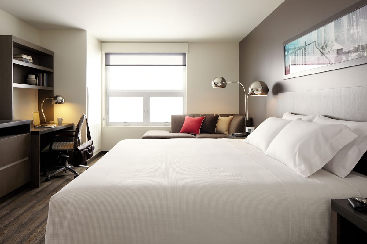 A hotel room with a large bed, a desk with a chair, a bookshelf, and a window providing natural light.
