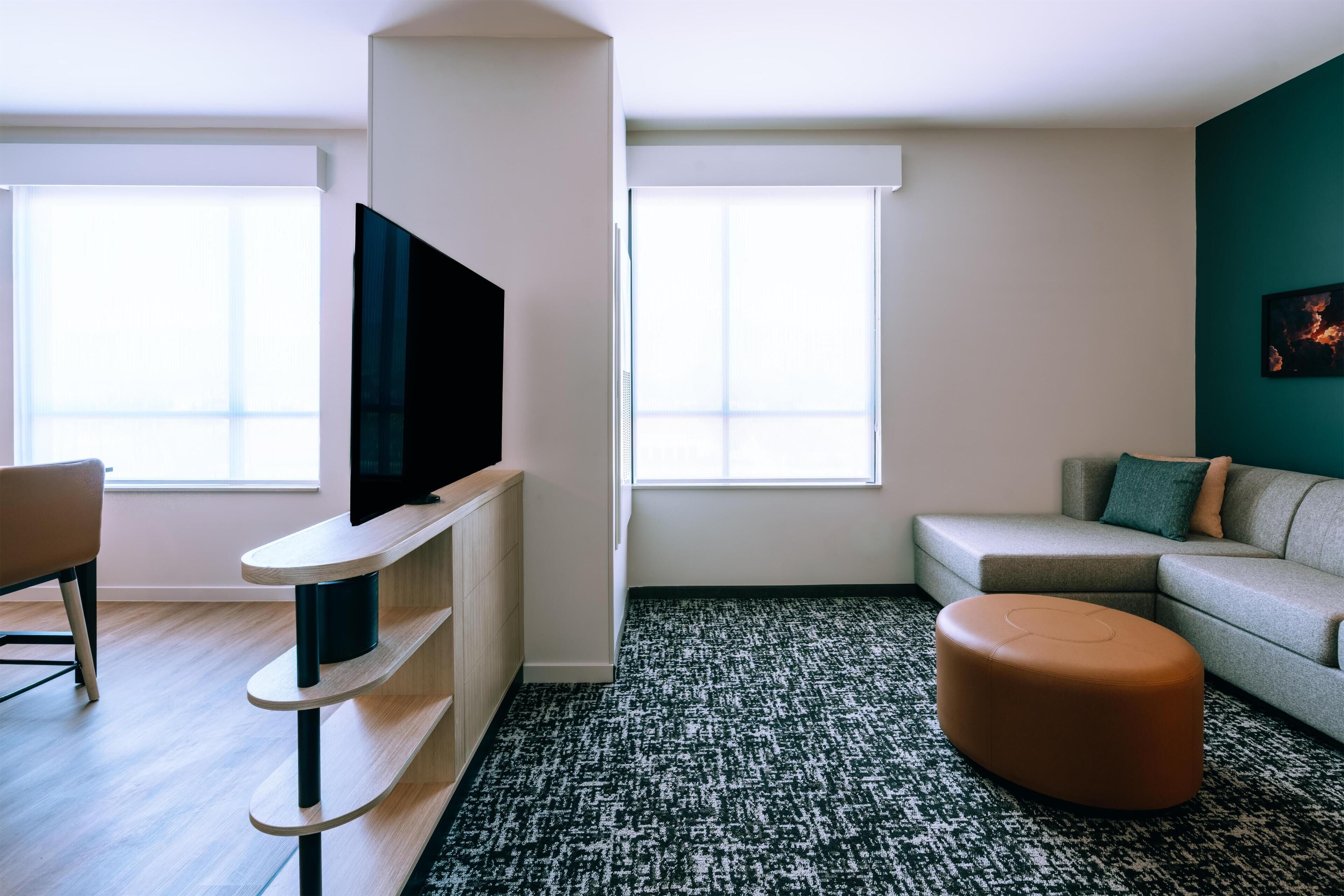 A modern living room with a flat-screen TV mounted on a wooden stand, a grey sofa with cushions, a brown ottoman, and a patterned rug.