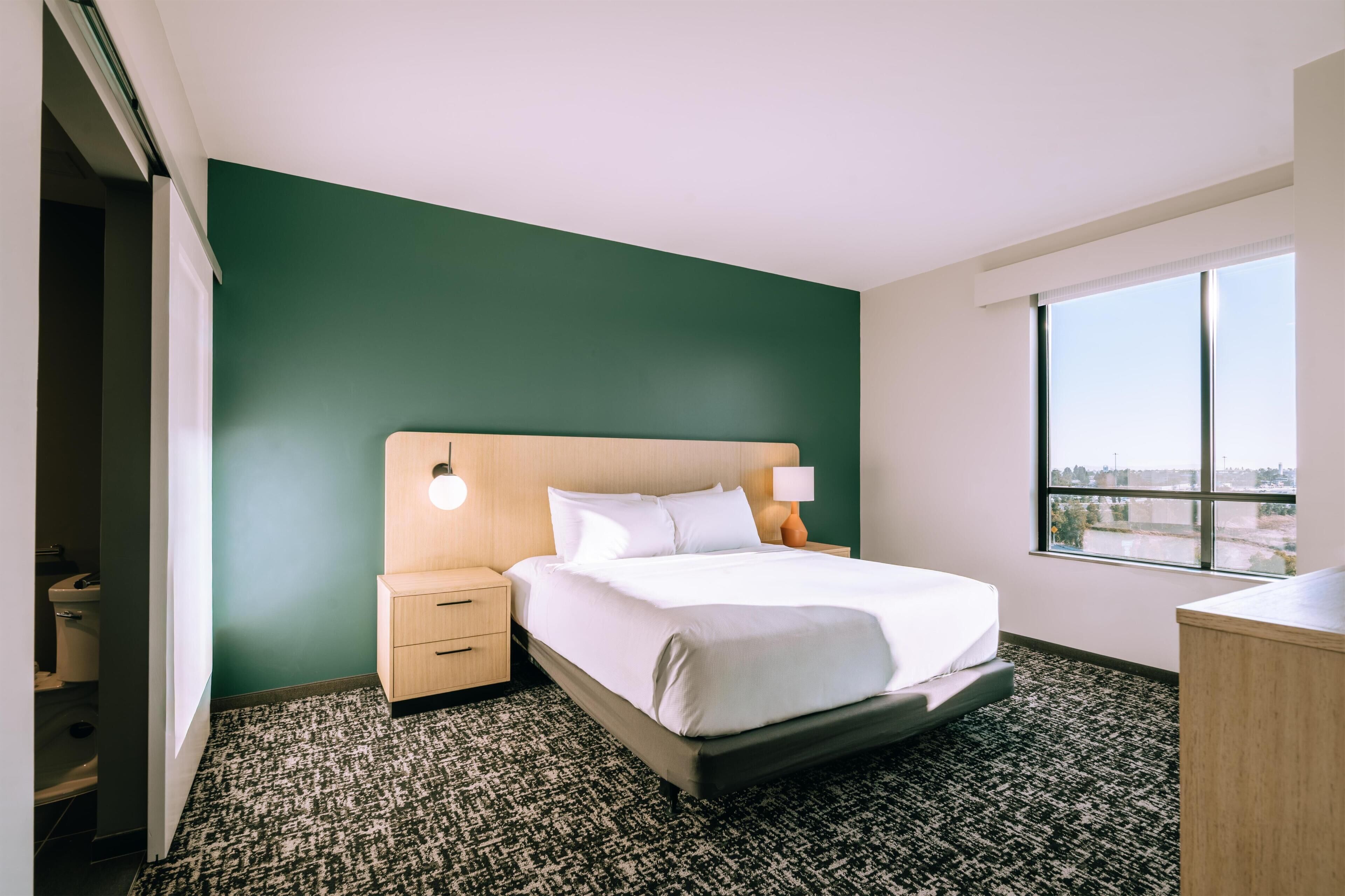 Habitación de hotel con cama, mesita de noche, lámpara, ventana con vistas y una pared verde.