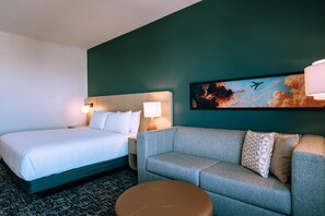 A hotel room with a bed, a sofa, a small round ottoman, a painting of clouds and an airplane, and a wall lamp. - Hyatt House Ontario Airport (Ontario)