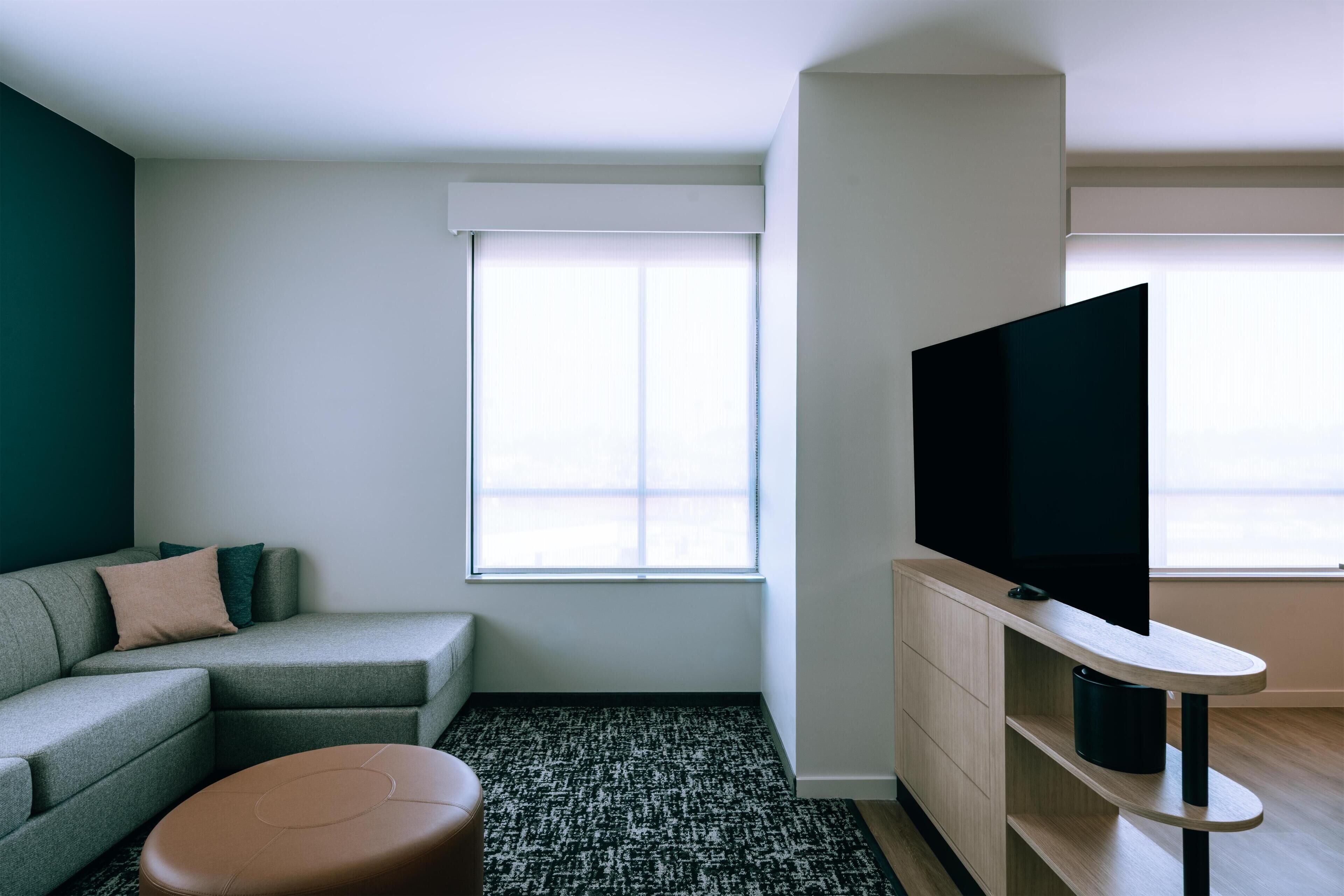 Una sala de estar moderna con un sofá gris, un reposapiés marrón, un televisor de pantalla plana sobre un soporte de madera y un ventanal con persianas.