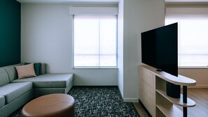 A modern living room with a grey sofa, a brown ottoman, a flat-screen TV on a wooden stand, and a large window with blinds.
