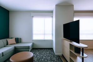 A modern living room with a grey sofa, a brown ottoman, a flat-screen TV on a wooden stand, and a large window with blinds. - Hyatt House Ontario Airport (Ontario)