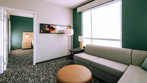 A hotel room with a sofa, ottoman, a painting, a lamp, and a view through a window. - Hyatt House Ontario Airport (Ontario)