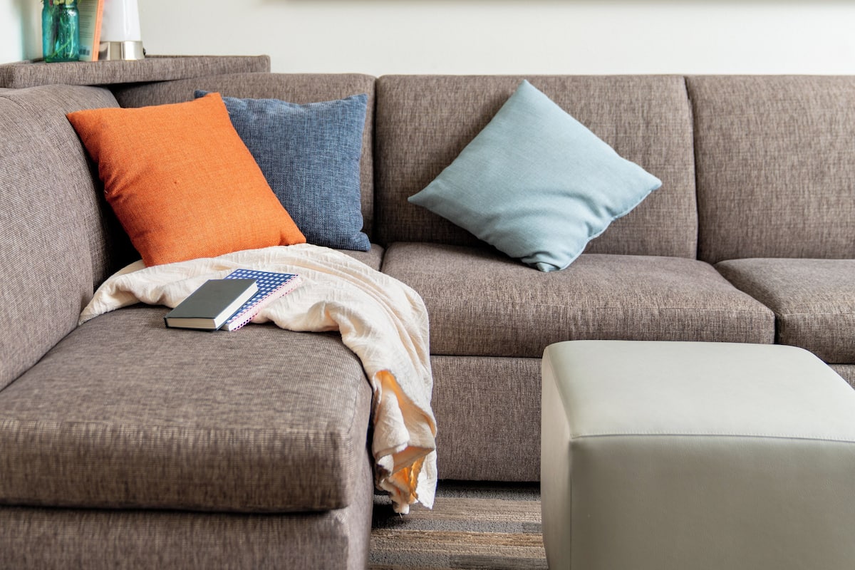 A living room with a grey sofa, two pillows, a blanket, and a book. A framed picture hangs on the wall above the sofa.