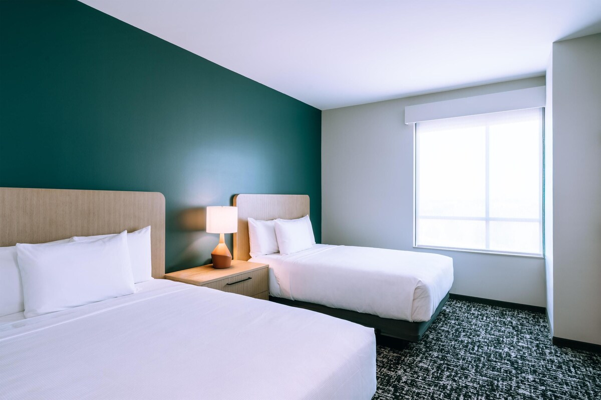 Ein Hotelzimmer mit zwei Betten, einem Nachttisch mit Lampe und einem großen Fenster, das für natürliches Licht sorgt.