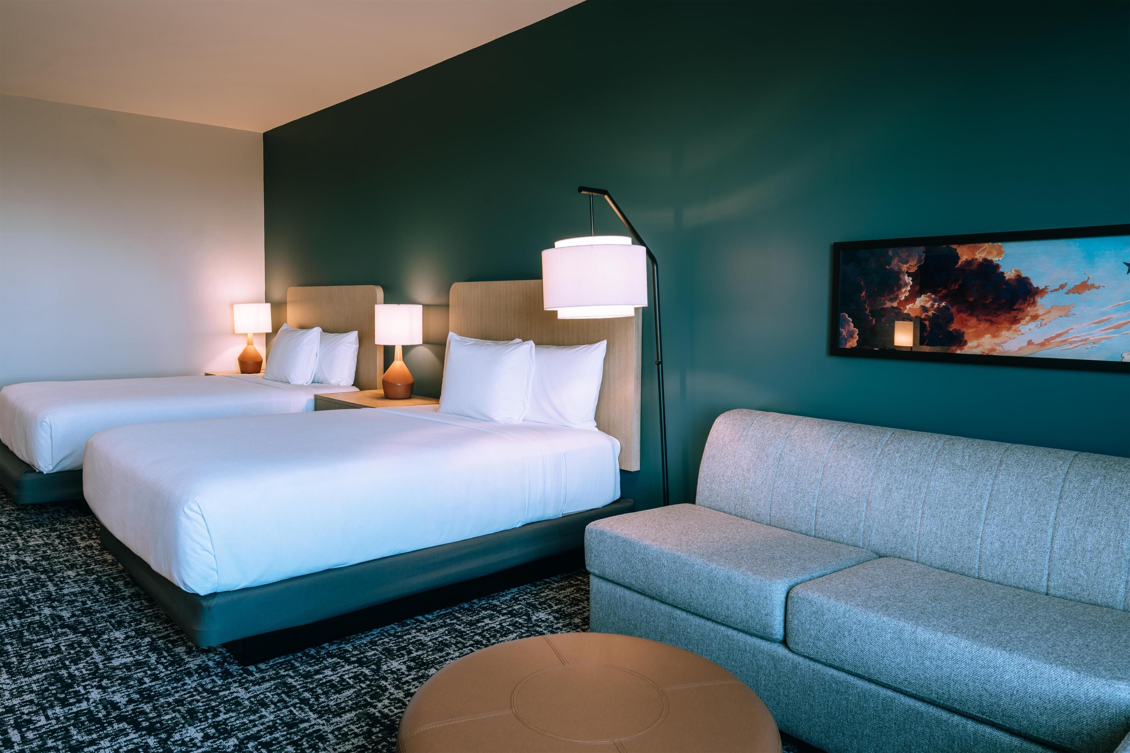 Habitación de hotel con dos camas, un sofá, una lámpara de pie y un cuadro en la pared.